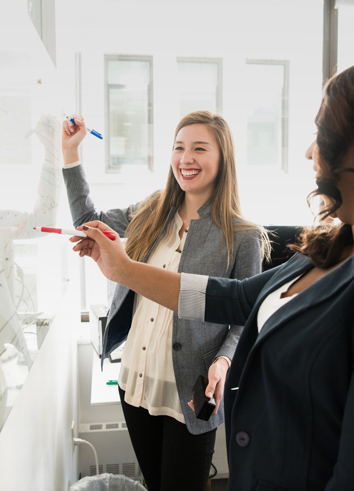 Two business women brainstorming on a whiteboard during a marketing planning workshop with their marketing agency